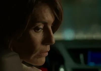 Close-up de mulher de cabelo castanho com olhar sério. Ela está em um carro escuro, à noite, com luzes de fundo. Clima misterioso e tenso.