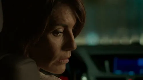 Close-up de mulher de cabelo castanho com olhar sério. Ela está em um carro escuro,à noite,com luzes de fundo. Clima misterioso e tenso.