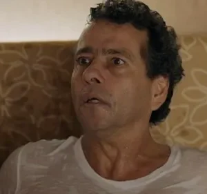 Homem suado, de meia-idade, com olhos arregalados e boca aberta, expressando choque ou medo. Usa camiseta branca.