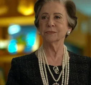 Uma mulher idosa elegante com cabelos grisalhos e pérolas. Ela olha séria em um fundo desfocado e colorido.