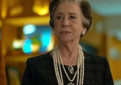 Uma mulher idosa elegante com cabelos grisalhos e pérolas. Ela olha séria em um fundo desfocado e colorido.