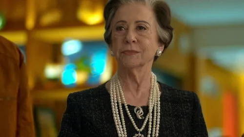 Babilônia – T01E112:Capítulo 112 1 Uma mulher idosa elegante com cabelos grisalhos e pérolas. Ela olha séria em um fundo desfocado e colorido.