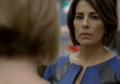 Mulher de cabelo escuro e vestido azul encara seriamente outra pessoa de costas, cabelo claro. Foco intenso e atmosfera dramática.