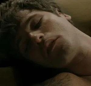 Close-up: homem de cabelo escuro, olhos fechados e boca entreaberta, dorme. Ombro nu com tatuagem em ambiente escuro e íntimo.