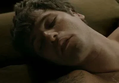 Close-up: homem de cabelo escuro, olhos fechados e boca entreaberta, dorme. Ombro nu com tatuagem em ambiente escuro e íntimo.