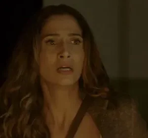 Mulher de cabelos longos, rosto assustado com olhos arregalados e boca aberta. Ambiente escuro e dramático.