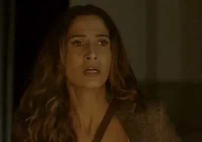 Mulher de cabelos longos, rosto assustado com olhos arregalados e boca aberta. Ambiente escuro e dramático.