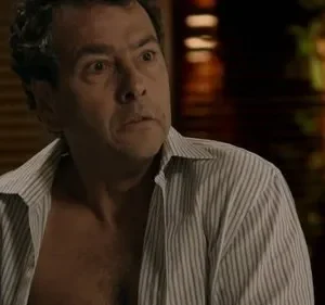 Homem de meia-idade com camisa aberta e expressão de surpresa ou choque. Olhos arregalados, boca ligeiramente aberta. Fundo escuro.