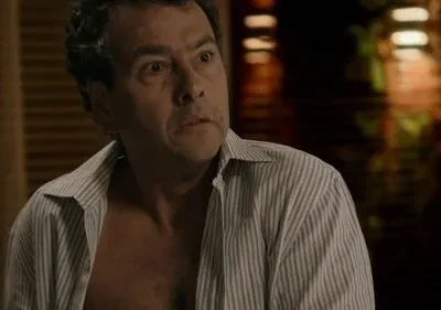 Homem de meia-idade com camisa aberta e expressão de surpresa ou choque. Olhos arregalados, boca ligeiramente aberta. Fundo escuro.