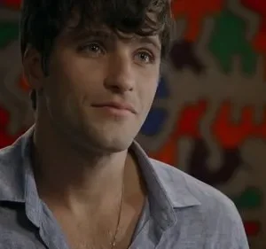 Close-up de um homem jovem com cabelo escuro e camisa azul, com um leve sorriso. Fundo desfocado com arte colorida abstrata.