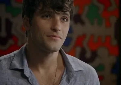 Close-up de um homem jovem com cabelo escuro e camisa azul, com um leve sorriso. Fundo desfocado com arte colorida abstrata.