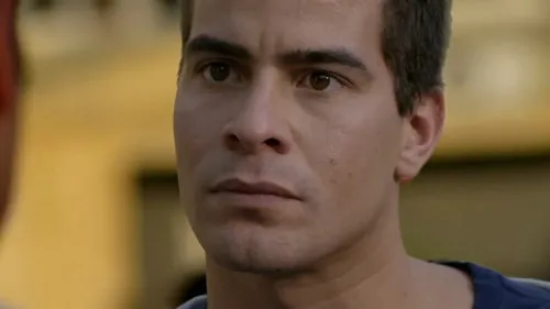 Close-up de homem sério com olhar intenso,encarando uma pessoa à esquerda. Fundo desfocado e quente,sugerindo tensão.