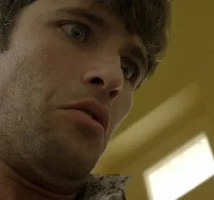 Rosto suado de homem com olhos arregalados e boca entreaberta, olhando para baixo. Fundo amarelo desfocado. Expressão de choque.