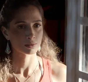 Mulher de cabelo cacheado e top laranja olha com expressão séria. Parte do rosto iluminada, com janela à direita.