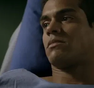 Homem jovem de pele morena, com expressão séria, em cama de hospital e avental azul. O ambiente é escuro.