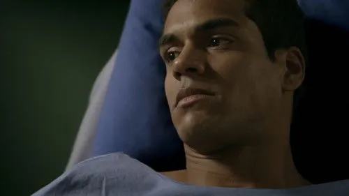 Homem jovem de pele morena,com expressão séria,em cama de hospital e avental azul. O ambiente é escuro.