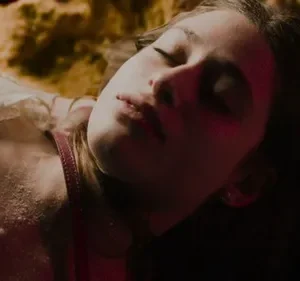 Close-up de jovem deitada, olhos fechados, com pele pálida e suja. Luz vermelha ilumina seu rosto. Cenário sombrio e texturizado.