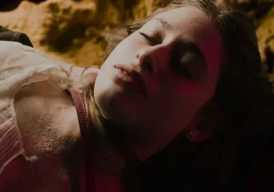 Close-up de jovem deitada, olhos fechados, com pele pálida e suja. Luz vermelha ilumina seu rosto. Cenário sombrio e texturizado.