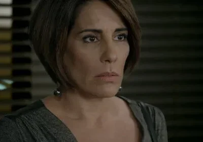 Close-up de mulher com cabelo castanho curto e semblante sério, usando blusa cinza e brincos, contra fundo escuro.