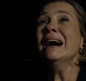 Close-up de mulher loira chorando intensamente, boca aberta, lágrimas visíveis. Fundo escuro realça o desespero.