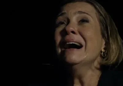 Close-up de mulher loira chorando intensamente, boca aberta, lágrimas visíveis. Fundo escuro realça o desespero.