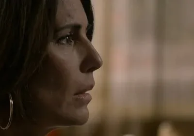 Mulher de perfil, cabelo escuro, argolas grandes, olhar sério e preocupado. Fundo desfocado com luz suave. Clima introspectivo.