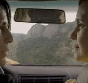 Duas mulheres em perfis num carro. Uma dirige, a outra é passageira. Montanhas verdes ao fundo. Clima introspectivo.