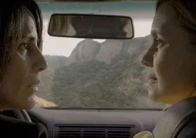 Duas mulheres em perfis num carro. Uma dirige, a outra é passageira. Montanhas verdes ao fundo. Clima introspectivo.