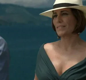 Uma mulher de chapéu e vestido verde olha para um homem de costas, com o mar azul ao fundo. Cenário costeiro e clima de verão.