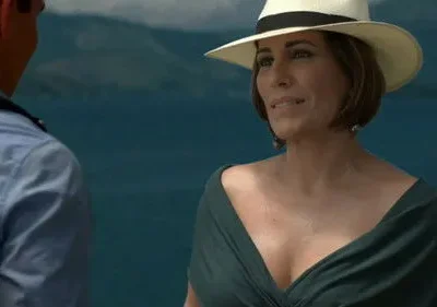 Uma mulher de chapéu e vestido verde olha para um homem de costas, com o mar azul ao fundo. Cenário costeiro e clima de verão.