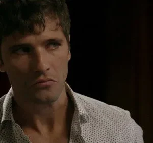 Homem de cabelo escuro e barba, camisa clara pontilhada, com expressão séria e pensativa. Fundo escuro e embaçado.