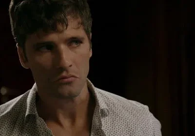 Homem de cabelo escuro e barba, camisa clara pontilhada, com expressão séria e pensativa. Fundo escuro e embaçado.