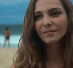 Mulher jovem com cabelo castanho claro sorri sutilmente. Ao fundo, praia, mar azul e salva-vidas desfocados.