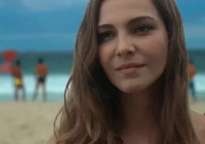Mulher jovem com cabelo castanho claro sorri sutilmente. Ao fundo, praia, mar azul e salva-vidas desfocados.