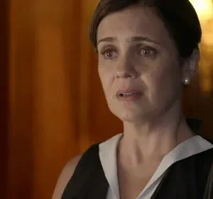 Close-up de mulher angustiada com olhos mareados, em blusa preta e branca. Fundo escuro com tons quentes. Clima de drama intenso.