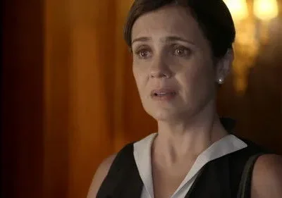 Close-up de mulher angustiada com olhos mareados, em blusa preta e branca. Fundo escuro com tons quentes. Clima de drama intenso.