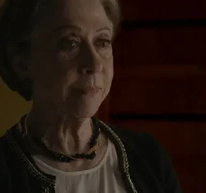 Mulher idosa de cabelo grisalho com olhar contemplativo. Veste casaco escuro, colar. Fundo sombrio e texturizado.