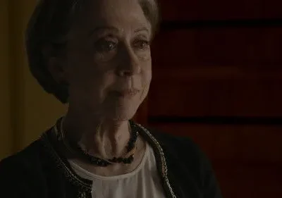 Mulher idosa de cabelo grisalho com olhar contemplativo. Veste casaco escuro, colar. Fundo sombrio e texturizado.