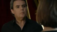 Homem grisalho de camisa preta olha sério para mulher de costas. Fundo escuro e dramático,clima tenso.