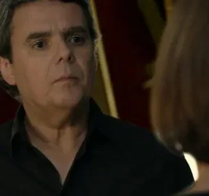 Homem grisalho de camisa preta olha sério para mulher de costas. Fundo escuro e dramático, clima tenso.