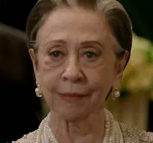 Close-up de mulher idosa elegante, cabelos grisalhos, joias de pérolas, sorrindo sutilmente. Fundo escuro e formal.