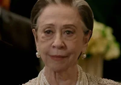 Close-up de mulher idosa elegante, cabelos grisalhos, joias de pérolas, sorrindo sutilmente. Fundo escuro e formal.