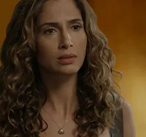 Close-up de mulher com cabelo cacheado castanho, colar de pérola. Expressão séria e introspectiva em fundo laranja-amarronzado.