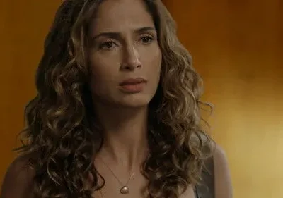 Close-up de mulher com cabelo cacheado castanho, colar de pérola. Expressão séria e introspectiva em fundo laranja-amarronzado.