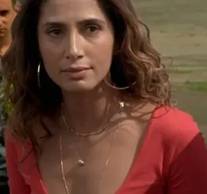 Mulher de top vermelho com expressão séria encara. Homem desfocado a observa atrás. Cenário externo com carro, clima tenso.