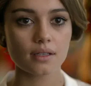 Close-up de mulher jovem de cabelo castanho curto, olhos escuros marcados e expressão pensativa. Fundo embaçado com tons quentes.