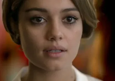 Close-up de mulher jovem de cabelo castanho curto, olhos escuros marcados e expressão pensativa. Fundo embaçado com tons quentes.