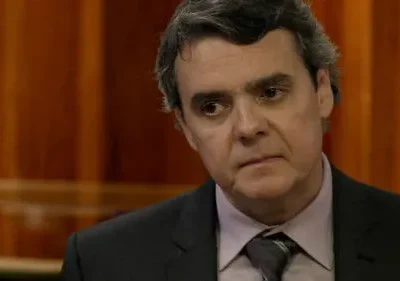 Homem de terno escuro e gravata, com cabelos grisalhos e expressão séria, olhando para o lado, em frente a painéis de madeira.