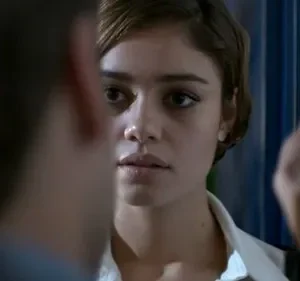 Mulher jovem de cabelo curto e camisa branca fita intensamente uma figura embaçada. Ambiente escuro com detalhes azuis, criando um clima tenso.