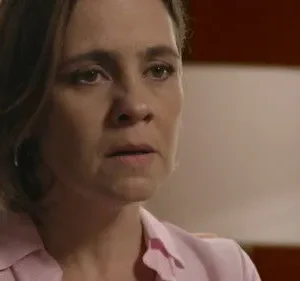 Close-up de mulher com cabelo castanho e camisa rosa, olhando com expressão séria e preocupada. Fundo interior desfocado.